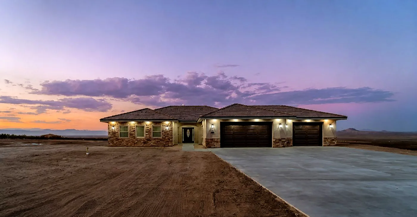 Custom home at twilight on open desert acreage, windows lit, mountains on the horizon